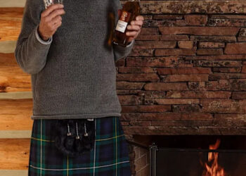 Man in a kilt holding a whiskey bottle and glass by a fireplace indoors.