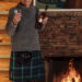 Man in a kilt holding a whiskey bottle and glass by a fireplace indoors.