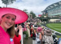 A smiling woman in a pink hat poses for a selfie at a crowded outdoor event with spectators in elegant hats.