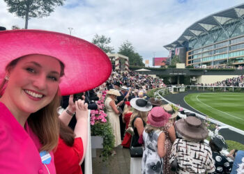 A smiling woman in a pink hat poses for a selfie at a crowded outdoor event with spectators in elegant hats.