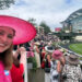 A smiling woman in a pink hat poses for a selfie at a crowded outdoor event with spectators in elegant hats.