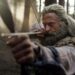 Hugh Jackman as a long-haired man drawing a bow and arrow in a forest.