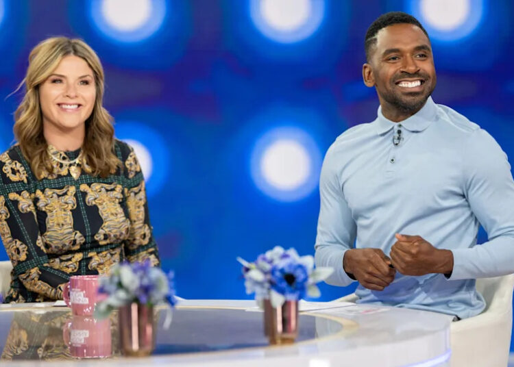 Two people seated at a table with blue flowers, smiling, in a studio with a blue, circular light background.