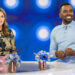 Two people seated at a table with blue flowers, smiling, in a studio with a blue, circular light background.