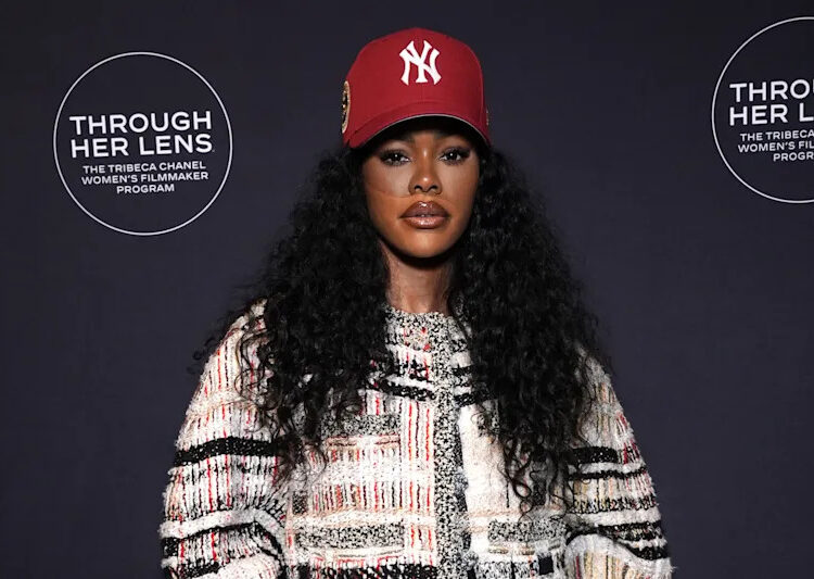 Teyana Taylor with long curly hair, wearing a red NY cap, patterned jacket, and brown pants, poses at the "Through Her Lens" event.