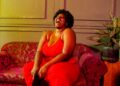 Full body of barefoot plump African American female laughing with mouth opened sitting on comfortable sofa