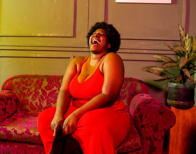 Full body of barefoot plump African American female laughing with mouth opened sitting on comfortable sofa