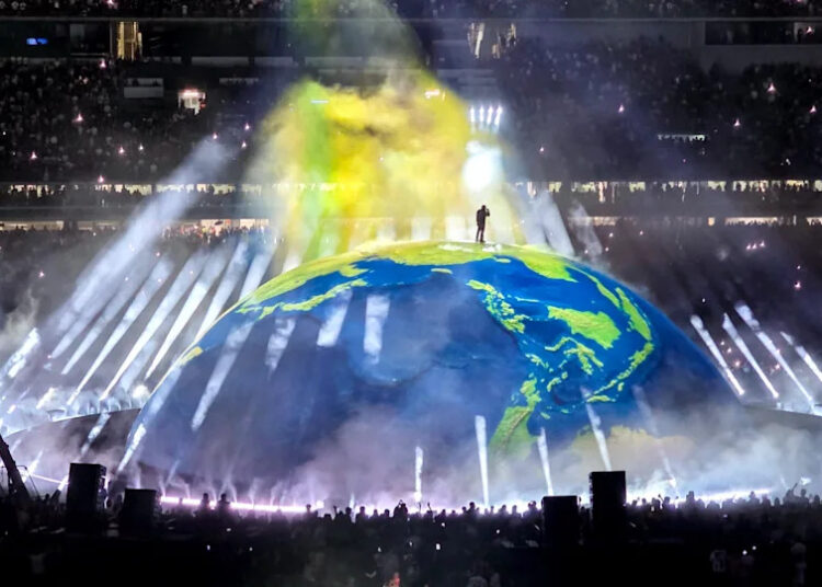 Ye stands on a large, illuminated globe surrounded by beams of light and smoke effects in a packed stadium.