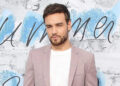 Liam Payne in a light pink blazer and white shirt, standing against a blue and white background.