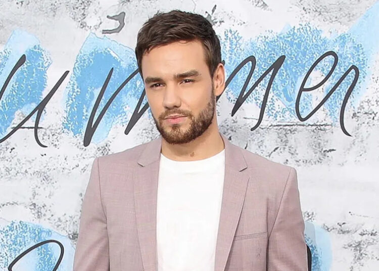 Liam Payne in a light pink blazer and white shirt, standing against a blue and white background.
