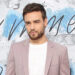 Liam Payne in a light pink blazer and white shirt, standing against a blue and white background.