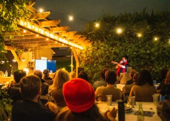 Photograph of a man standing in front of a seated audience outdoors at night indicating to a video screen and holding a microphone.
