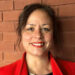 Michelle Daurat has brown hair and eyes and her hair is tied back. She is wearing a red lipstick, which matches her coat, and a black top. She is smiling.
