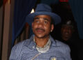 Max B in a blue hat and checkered shirt walks on a red carpet. He's wearing jeans with black designs and accessorized with jewelry.
