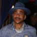 Max B in a blue hat and checkered shirt walks on a red carpet. He's wearing jeans with black designs and accessorized with jewelry.