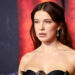 Millie Bobby Brown at an event, wearing a strapless black dress with drop earrings, against a red and black background.