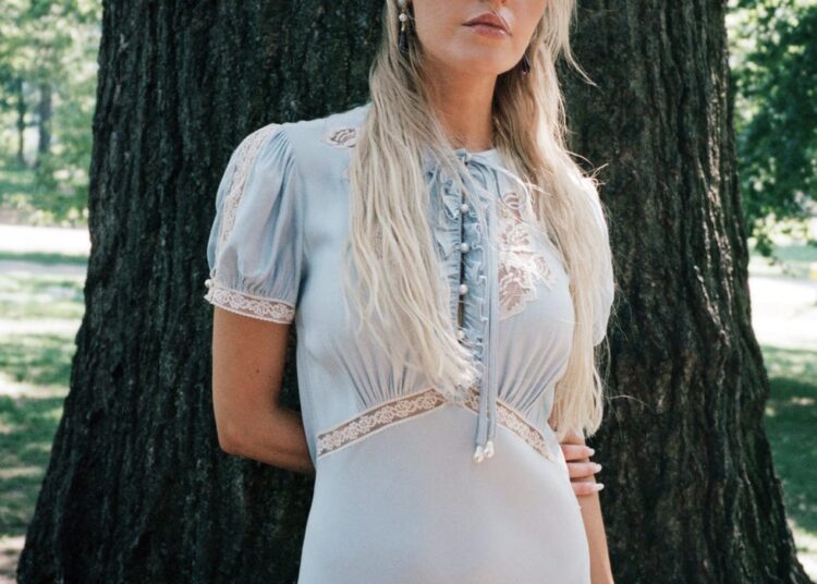 Morgan in a light blue dress stands against a tree in a park.