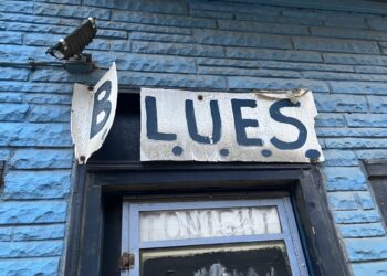 Sign that reads "B.L.U.E.S." over window opening on side of brick building.