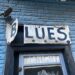 Sign that reads "B.L.U.E.S." over window opening on side of brick building.