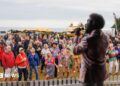 A man on stage with black hair and a pink satin jacket sings into a microphone as he looks out into a crowd of people. Behind the audience is a row of stalls and fairground rides and beyond that is the sea.
