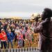 A man on stage with black hair and a pink satin jacket sings into a microphone as he looks out into a crowd of people. Behind the audience is a row of stalls and fairground rides and beyond that is the sea.