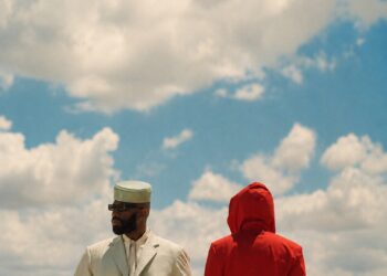 Two figures, one in a white suit and one in a red hooded jacket, stand against a cloudy sky.