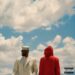 Two figures, one in a white suit and one in a red hooded jacket, stand against a cloudy sky.