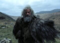 A gray-haired and heavily bearded Hugh Jackman wears a woolly coat while standing against a hilly backdrop.