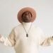Nick Cooper wears a cream turtleneck sweater and matching wide-brim hat while looking upward with both arms raised during a portrait session against a white background.