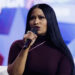 Nicki Minaj with long black hair speaks into a microphone on stage, wearing a dark outfit. The background is blurred.