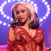 Cardi B performing on stage in a red, embellished outfit with dramatic lighting and a halo of lights in the background.