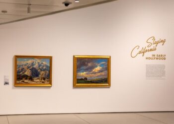 Two paintings, one of mountains and one of a field below a graying sky, hang on a white wall.