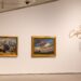 Two paintings, one of mountains and one of a field below a graying sky, hang on a white wall.