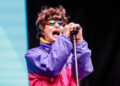 Singer passionately performing on stage, wearing a colorful jacket and reflective sunglasses while holding a microphone