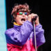 Singer passionately performing on stage, wearing a colorful jacket and reflective sunglasses while holding a microphone