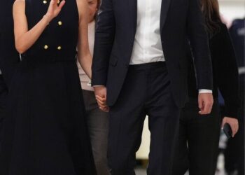 An individual in formal attire walks alongside another person dressed in a black sleeveless top, both of them appearing to be en