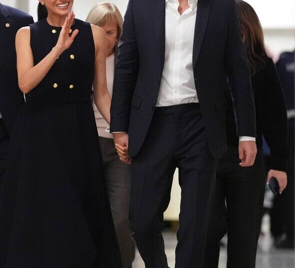 An individual in formal attire walks alongside another person dressed in a black sleeveless top, both of them appearing to be en