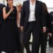 An individual in formal attire walks alongside another person dressed in a black sleeveless top, both of them appearing to be en