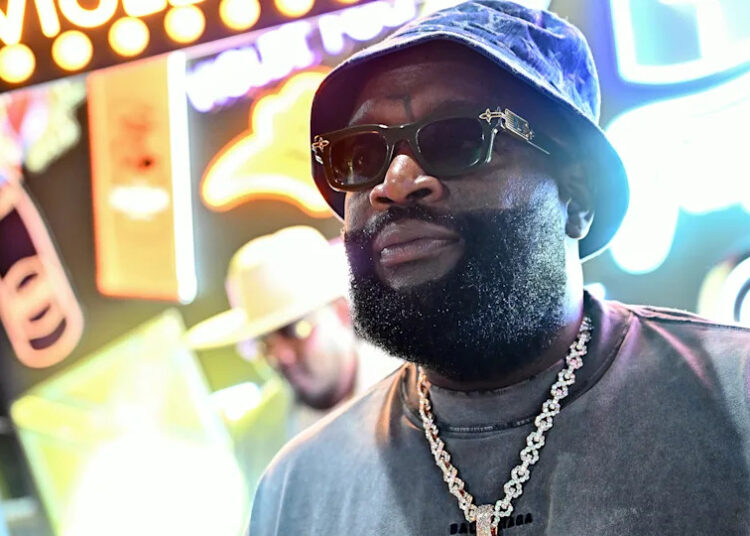 Rick Ross, wearing sunglasses and a blue bucket hat, stands in front of colorful neon signs, with another person blurred in the background.
