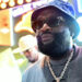 Rick Ross, wearing sunglasses and a blue bucket hat, stands in front of colorful neon signs, with another person blurred in the background.
