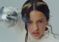 close-up photo of singer Rosalía with a halo-shaped highlight in her hair, pointing a sword into the camera