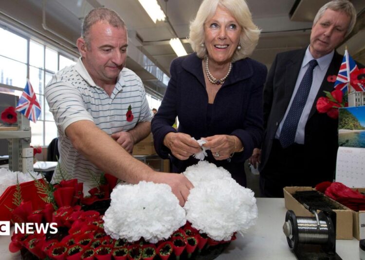Man in striped shirt holds white flowers on top of red poppy wreath as woman with pearls smiles and prepares to add flowers