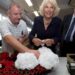 Man in striped shirt holds white flowers on top of red poppy wreath as woman with pearls smiles and prepares to add flowers