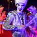 Drummers in metallic costumes with glowing drums and face paint performing in parade.
