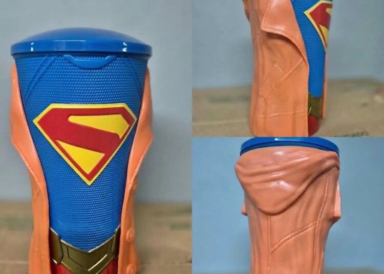 A Supergirl movie drink cup with a blue cup, Superman logo, gold belt buckle, and orange jacket.