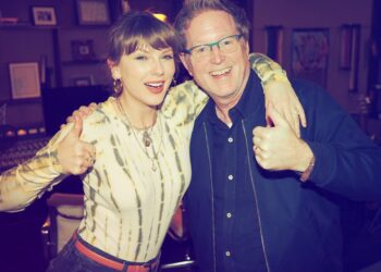 Taylor Swift wearing a tie-dye Agolde bodysuit with black jeans and opal jewelry with brown bangs and a ponytail posing in a recording studio