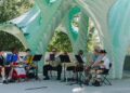A small band in traditional Bavarian attire performs outdoors under a large, modern, turquoise and white sculptural canopy, with trees and sunlight in the background.