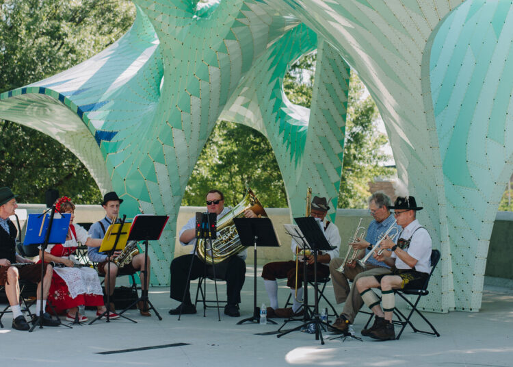 A small band in traditional Bavarian attire performs outdoors under a large, modern, turquoise and white sculptural canopy, with trees and sunlight in the background.