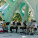 A small band in traditional Bavarian attire performs outdoors under a large, modern, turquoise and white sculptural canopy, with trees and sunlight in the background.