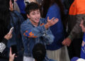Timothée Chalamet in a denim jacket with orange accents gestures animatedly in a crowd at a game.